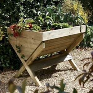 The Forest Kitchen Garden Trough 3x3, full of plants, situated on gravel, surrounded by shrubs.